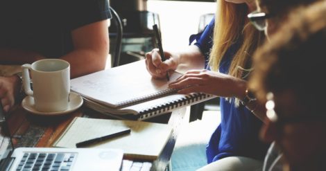 Woman writing on notebook at table with other people.