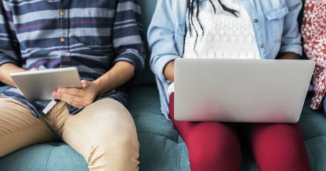 Two people sitting next to each other working on their tablet and laptop.